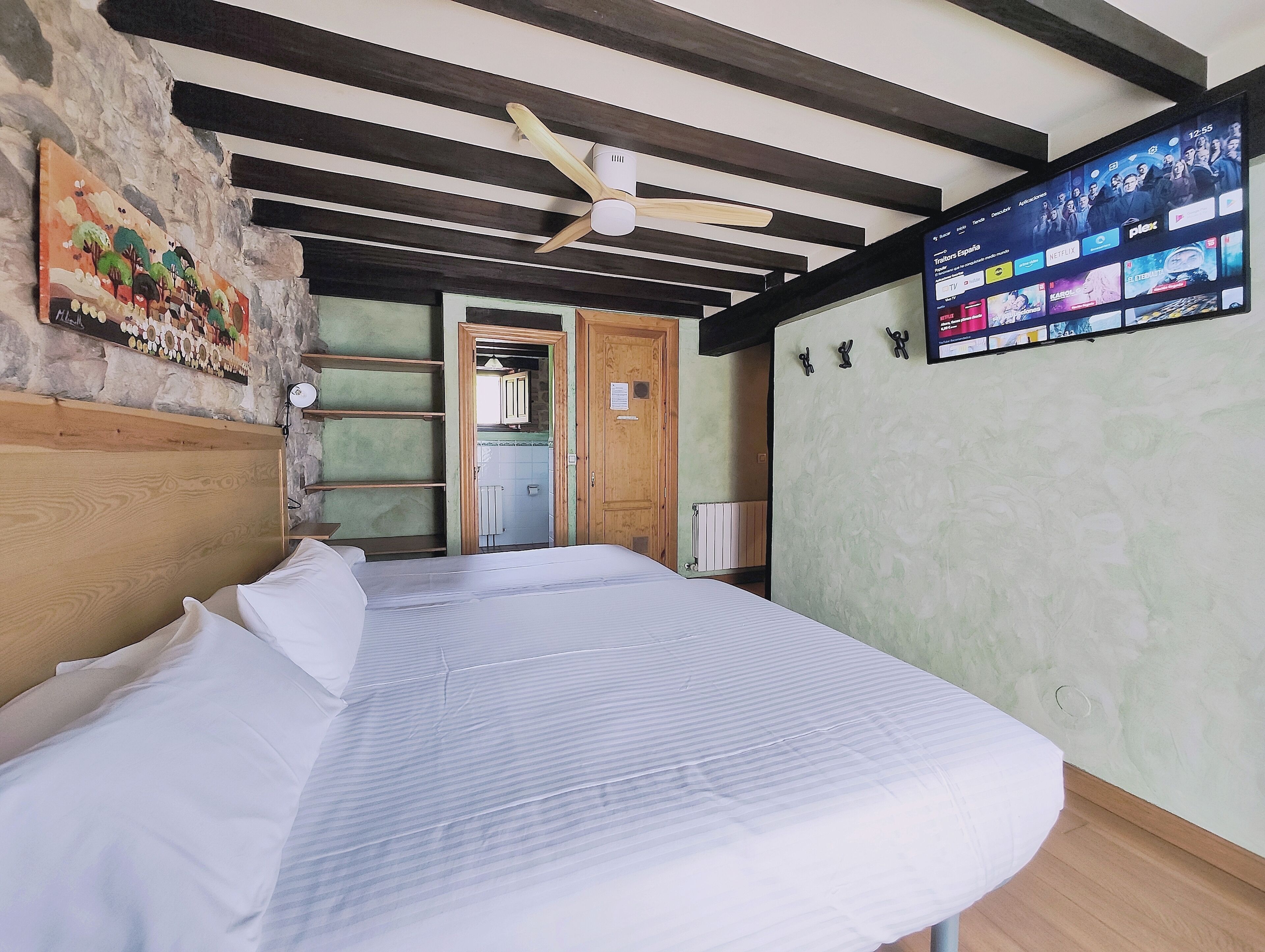 Een slaapkamer met een bed, houten hoofdbord, een plafondventilator, een aan de muur bevestigde televisie waarop een streamingdienst wordt afgespeeld, en een stenen muur met een schilderij.