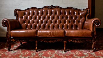 A brown leather sofa with a high backrest and ornate carvings, positioned on a red patterned carpet in a room with a white brick wall adorned with framed photographs.