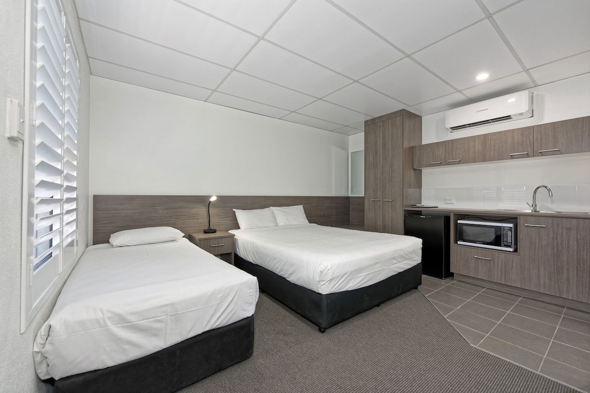 A hotel room with two beds, a desk, a microwave, and a kitchenette.