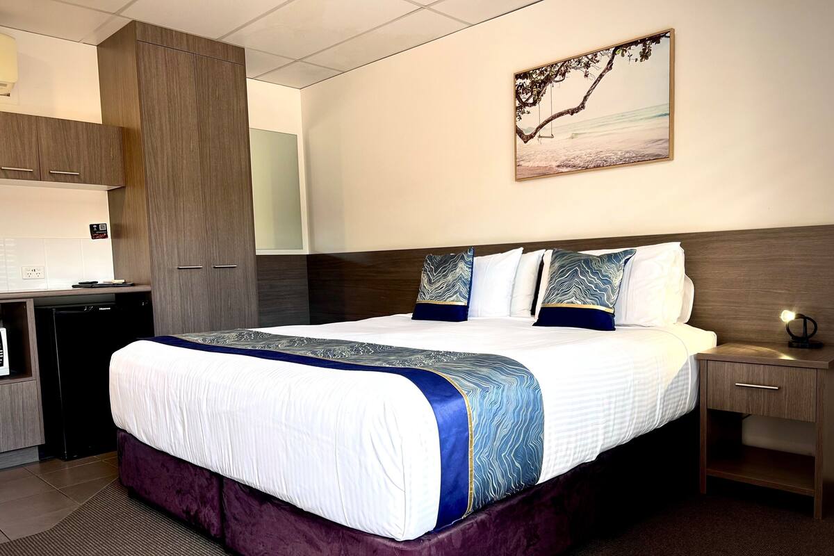 Quarto de hotel com uma cama grande, uma mesa de cabeceira, uma escrivaninha com um computador e uma cozinha.