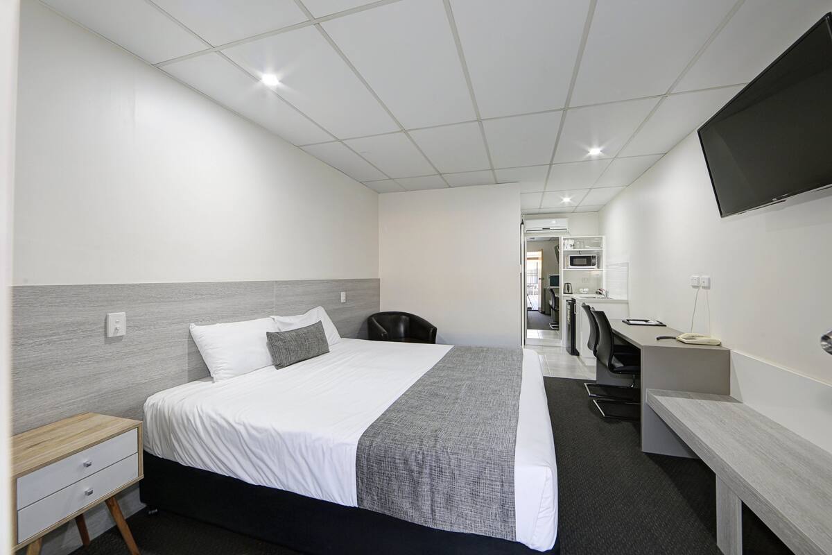 Quarto de hotel com cama, escrivaninha, cadeira, TV e uma cozinhazinha ao fundo.