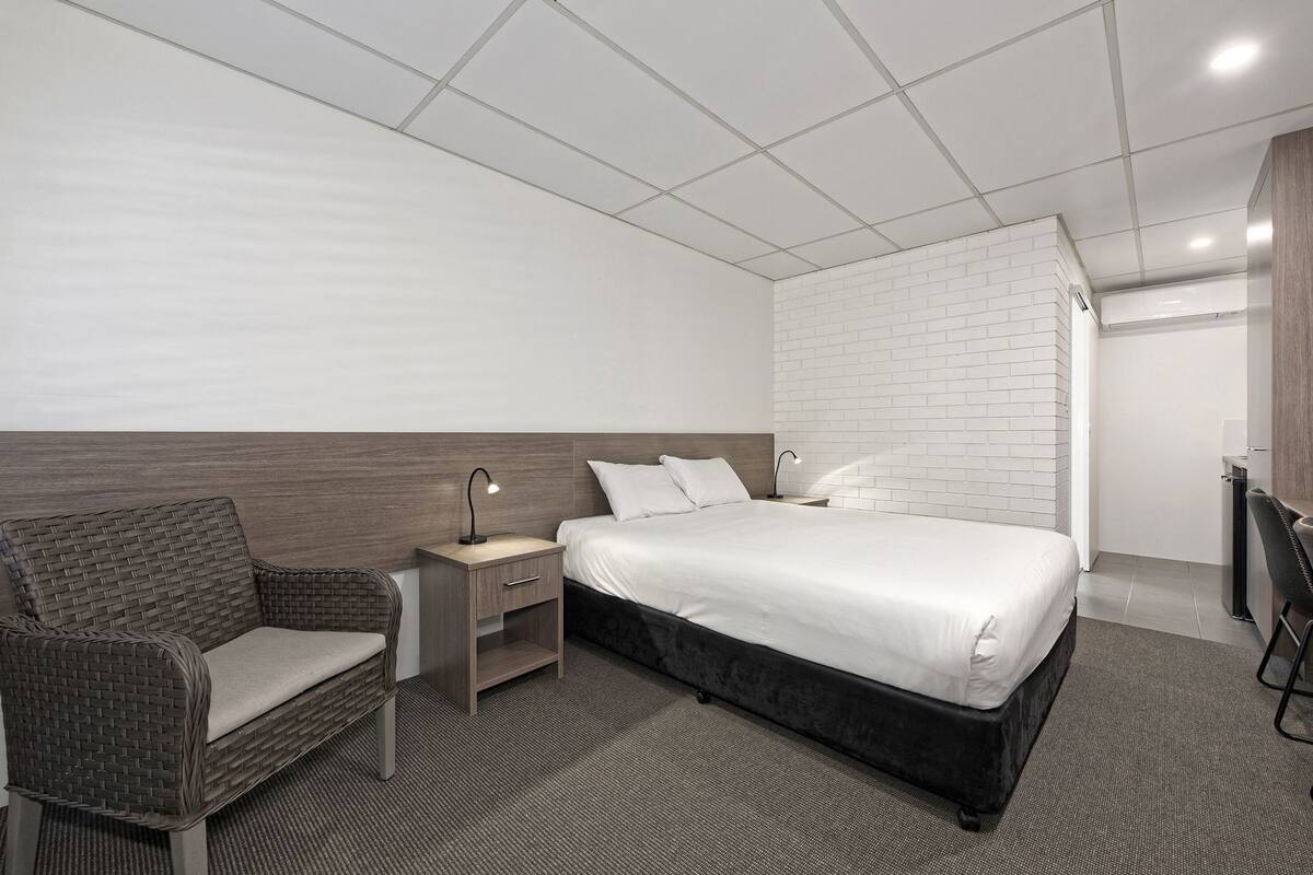 Quarto de hotel com uma cama, mesa de cabeceira, cadeira, escrivaninha e parede de tijolos brancos.