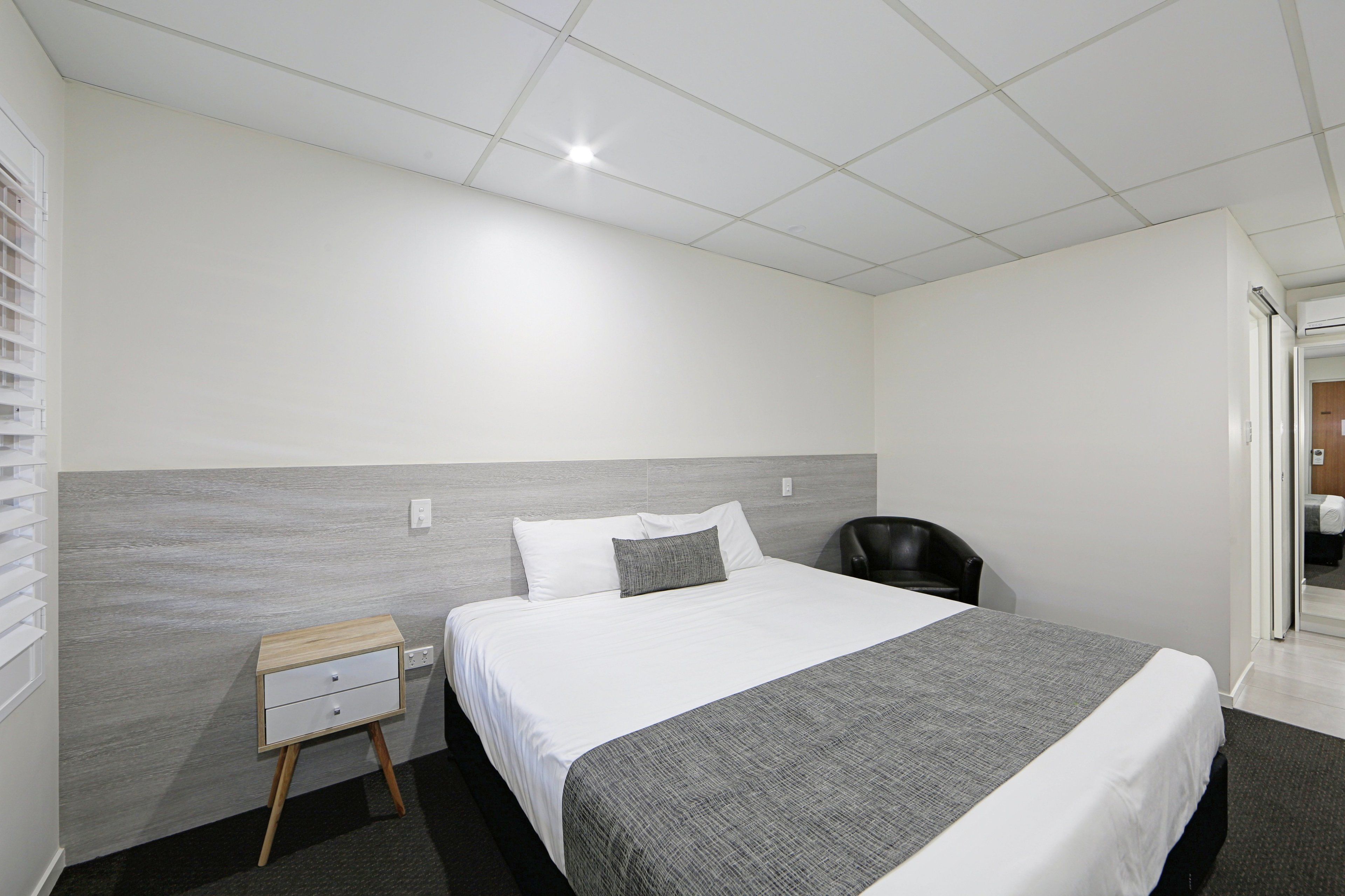 A hotel room with a large bed, a nightstand, a chair, and a view of the hallway.