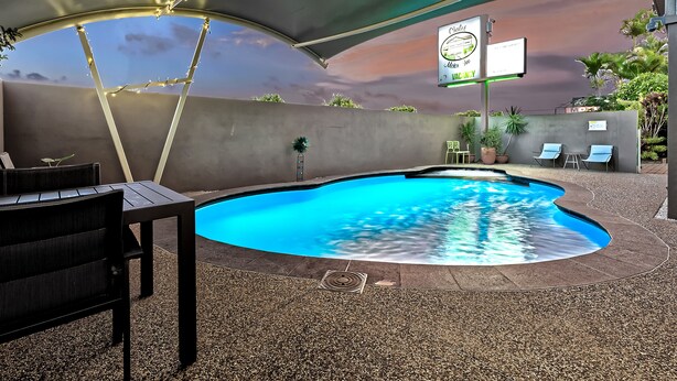 Una piscina de borde curvo, rodeada por una zona techada con una toldo. Hay sillas de descanso y una mesa con sillas.