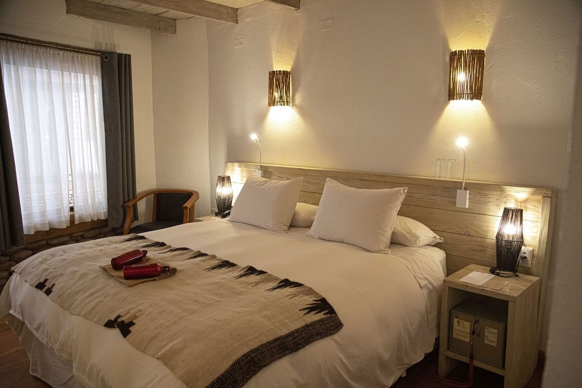 Ein Hotelzimmer mit einem großen Bett, zwei Nachttischlampen, einem Nachttisch mit Lampe, einem Sessel und einem Fenster mit Vorhängen.