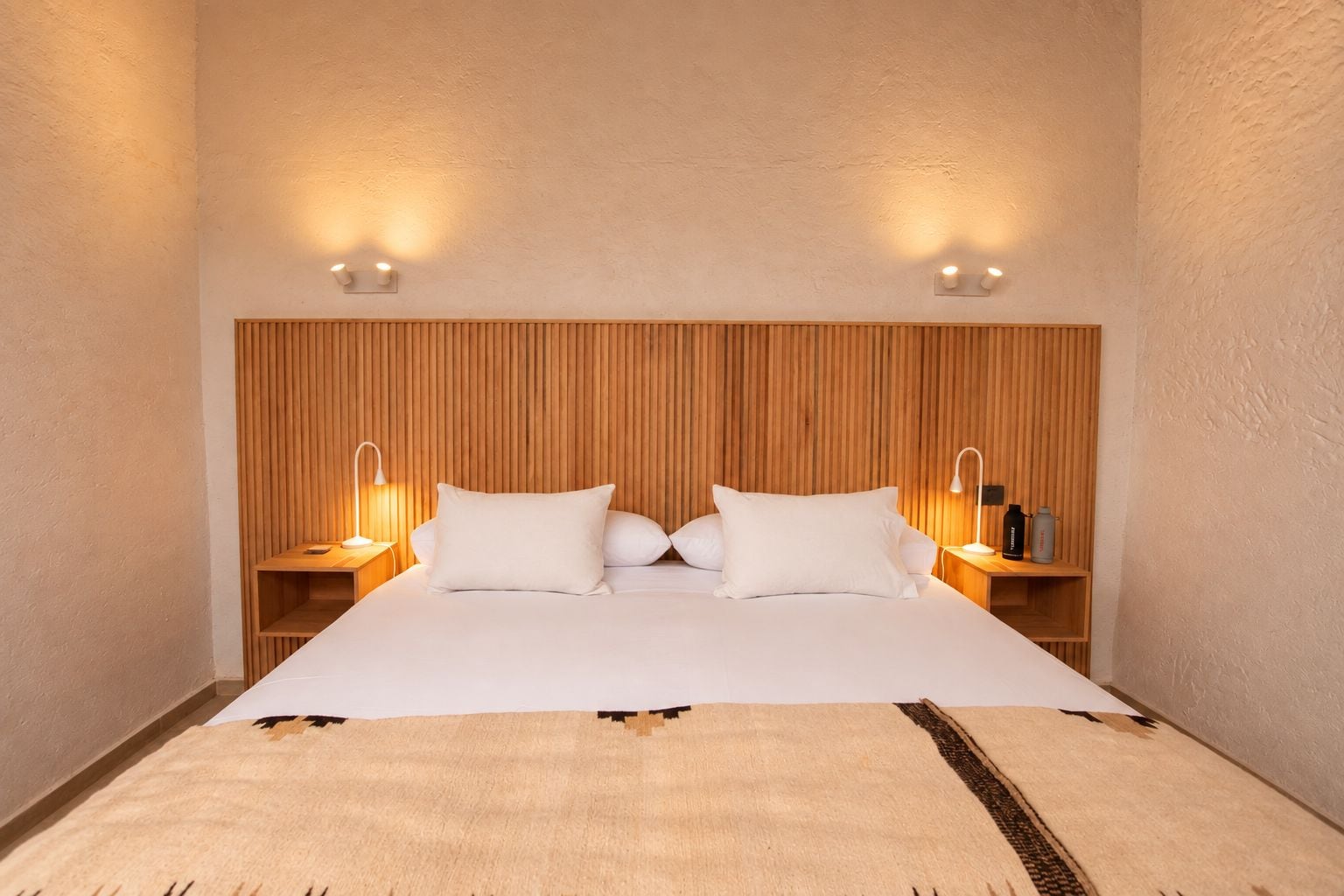 Habitación de hotel con una cama grande, cabecera de madera, dos mesitas de noche con lámparas y una lámpara de pared.