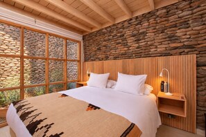 Slaapkamer met een stenen muur, houten balken in het plafond, een groot bed, nachtkastjes en een raam met uitzicht op beplanting.