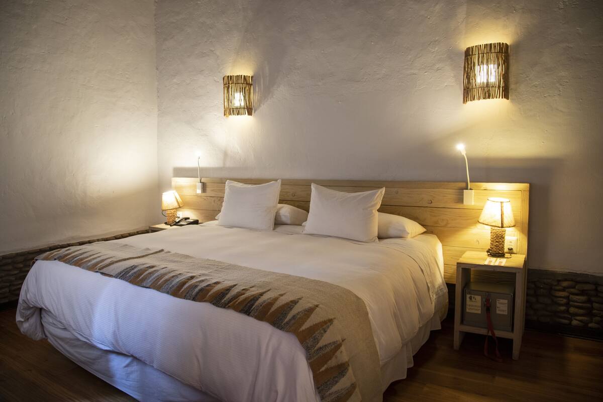 Ein ordentlich bezogenes Bett mit weißen Leinen und einer gemusterten Tagesdecke, zwei Nachttischlampen und ein Holz-Kopfstück.