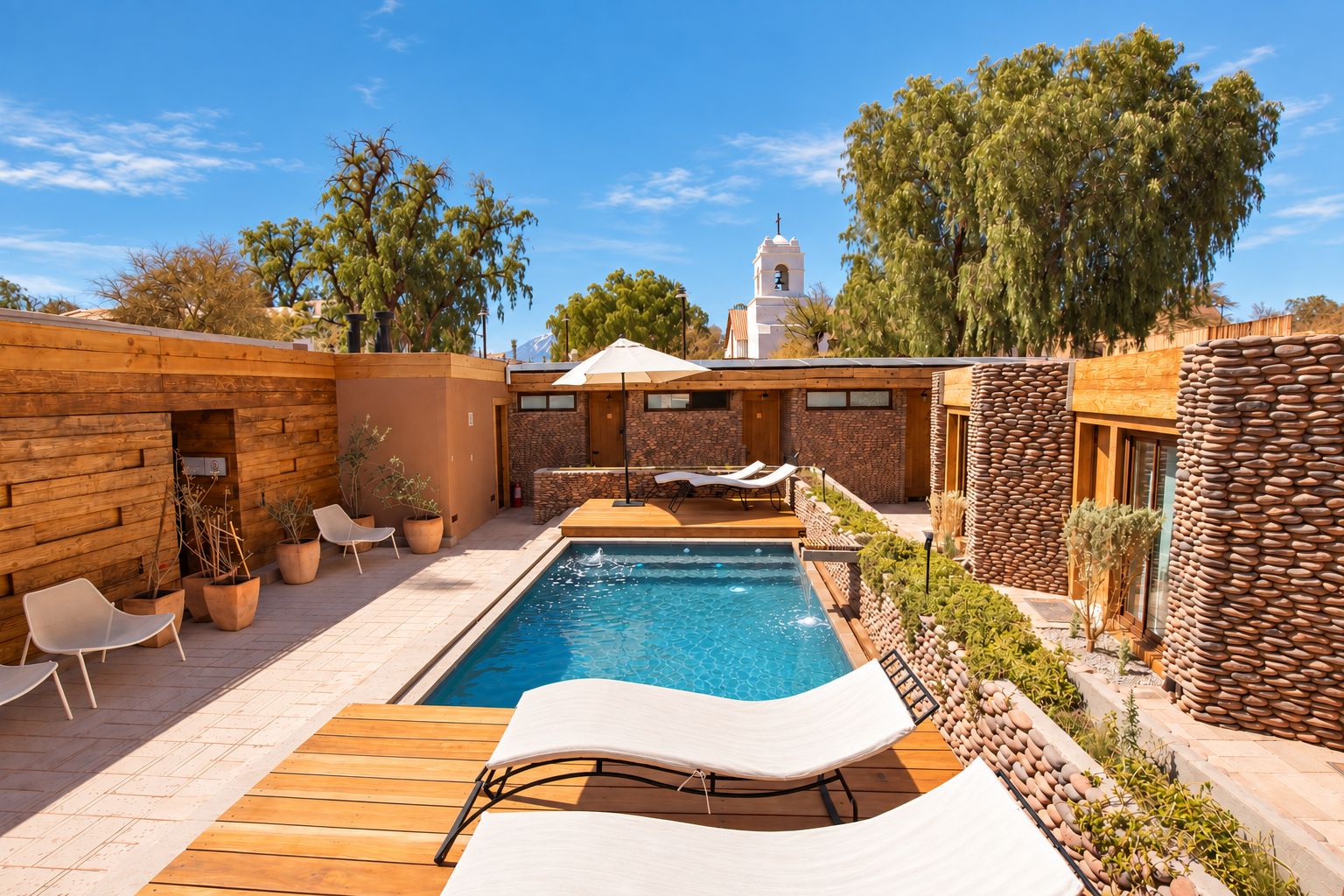 Un área de piscina con sillas de descanso, un muro de piedra y una torre campanario al fondo.
