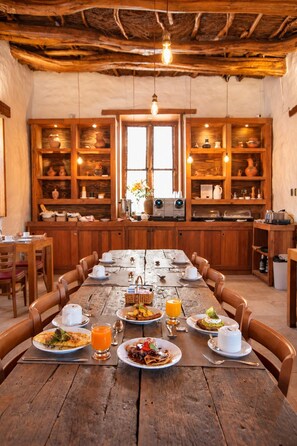 Un coin repas rustique, avec une longue table en bois dressée pour le petit-déjeuner, entouré d’armoires en bois dotées d’étagères.