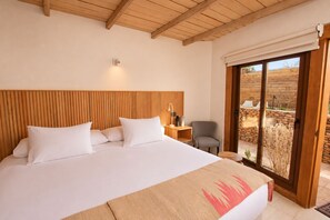 Een slaapkamer met een groot bed, een houten hoofdeinde, een nachtkastje met een lamp en uitzicht op een stenen muur door een glazen schuifdeur.