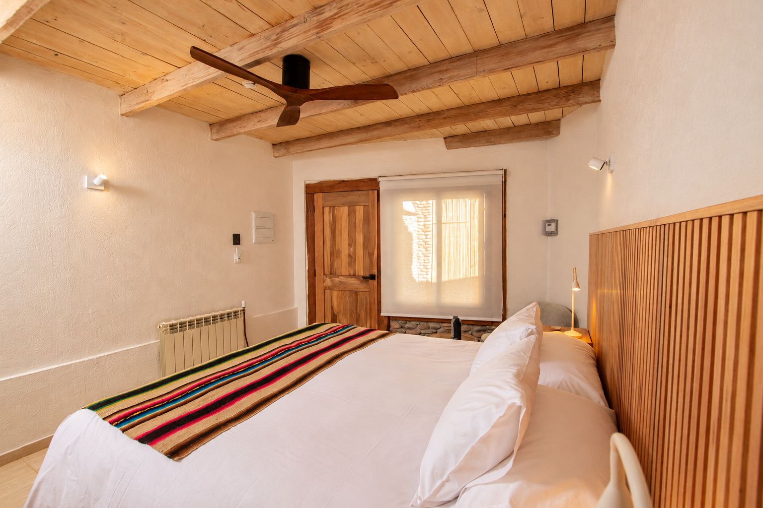 Un dormitorio con cama, cabecero de madera, ventilador de techo, ventana con cortinas y radiador.