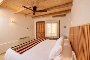 Een slaapkamer met een bed, houten hoofdbord, een plafondventilator, een raam met gordijnen en een radiator.