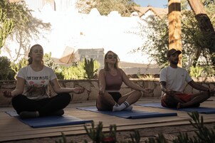 Tres personas meditando sobre esterillas de yoga al aire libre, con un muro de piedra y árboles en el fondo.