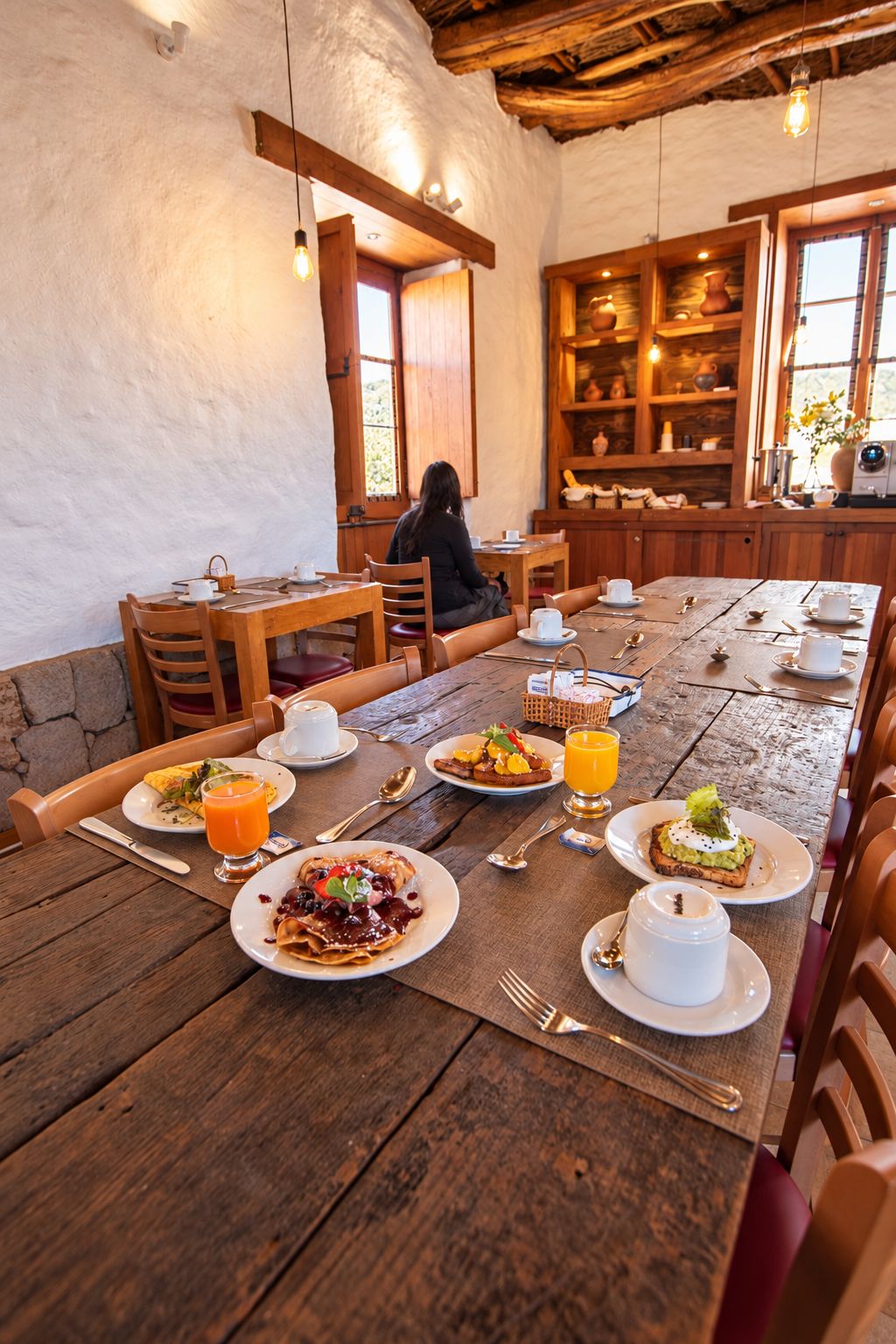 A rustic dining area with wooden tables and chairs, a person seated at a table, and a kitchen area with wooden cabinets and a window.