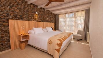 A bedroom with a stone wall, a wooden headboard, a bed with white bedding, a bedside table with a lamp, a chair, and a large window with curtains.