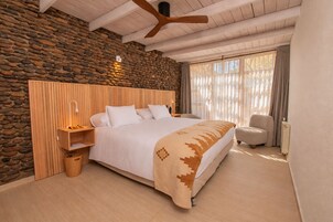 Un dormitorio con pared de piedra, cabecera de madera, cama con ropa de cama blanca, mesita de noche con lámpara, una silla y un ventanal con cortinas.