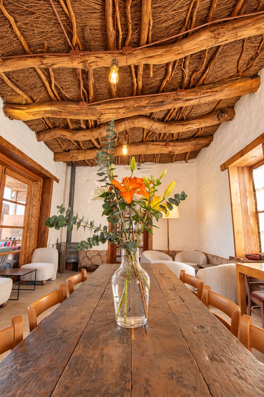 A rustic interior with a wooden table, chairs, a vase with flowers, and a thatched ceiling.