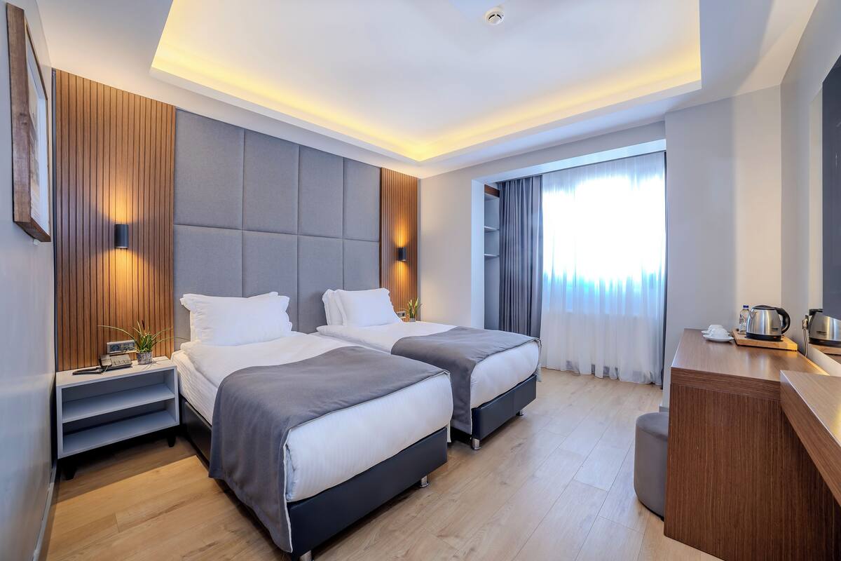 A modern hotel room with two beds, a wooden accent wall, a built-in desk, and a large window with curtains.