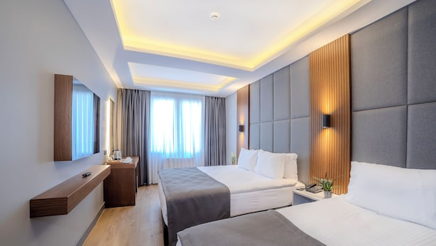 A modern hotel room with two beds, a wooden bedside table, a built-in TV, and a large window with curtains.