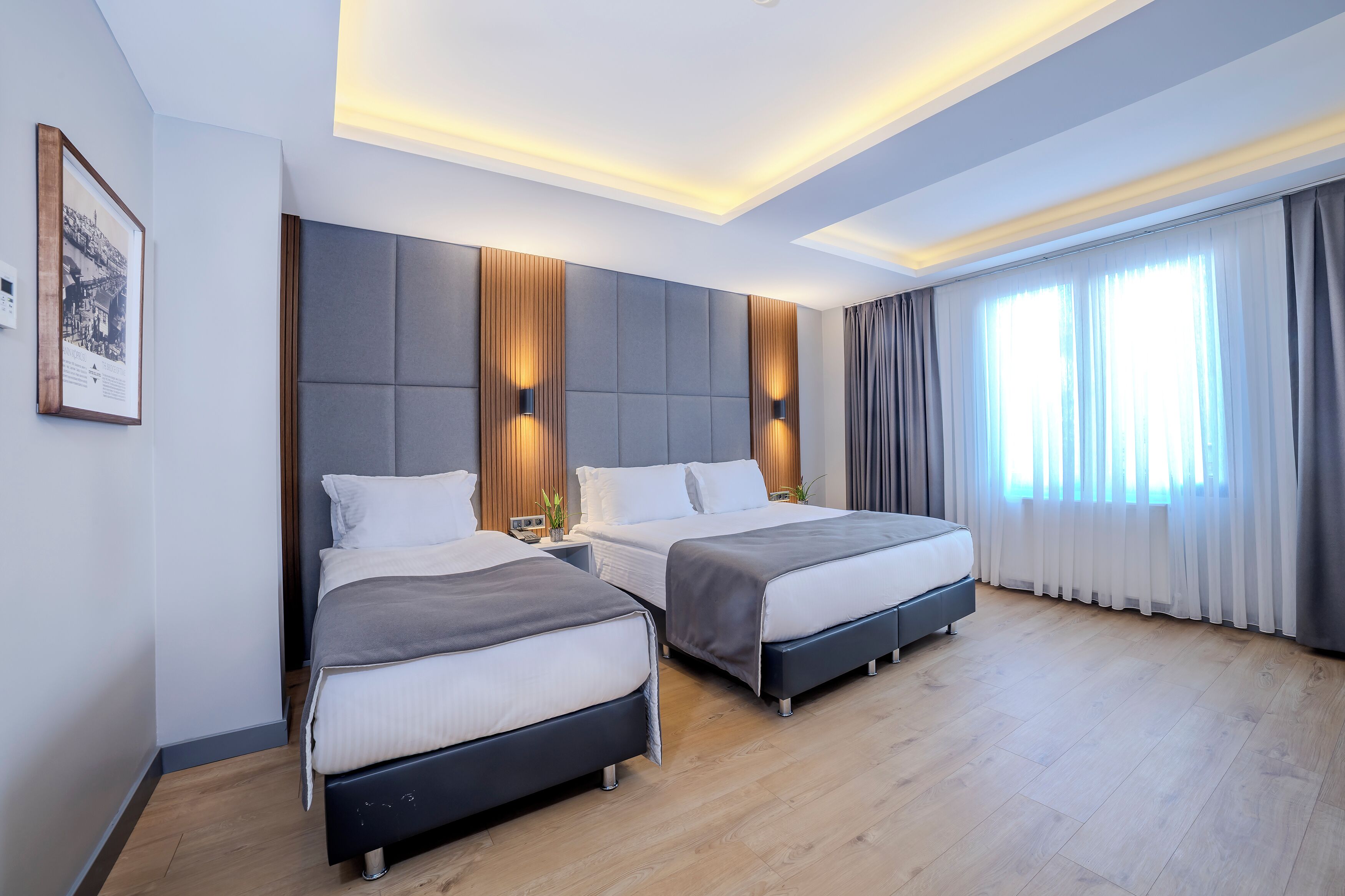 A modern hotel room with two beds, a large window with curtains, and a framed picture on the wall.