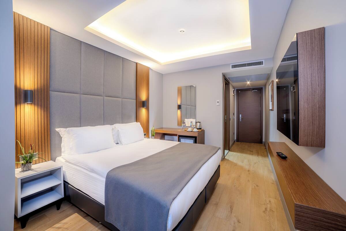 A modern hotel room with a large bed, bedside tables, a desk, a mirror, and a hallway leading to a door.