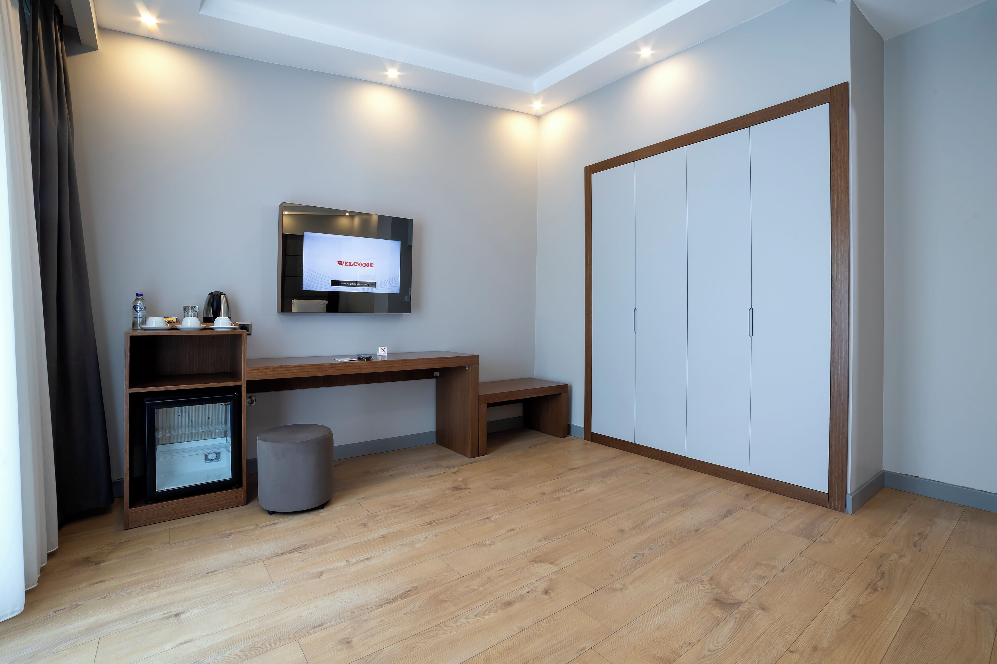 A modern hotel room with a wooden desk, a television mounted on the wall, a wardrobe, and a small trash bin.