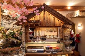 A rustic wooden structure with a glass display case containing food items, surrounded by a decorative rock garden and pink flowers. - Eder Michaela (Saalbach-Hinterglemm)