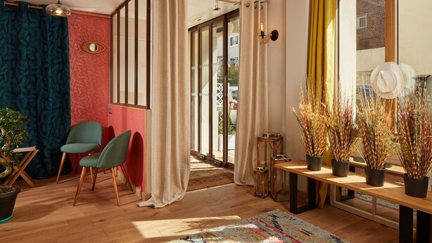 A modern living room with a wooden floor, a colorful rug, a green chair, a wooden table with decorative items, and a large window with curtains. - Hôtel Villa Sorel (Boulogne-Billancourt)