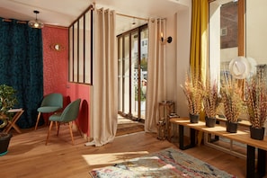 A modern living room with a wooden floor, a colorful rug, a green chair, a wooden table with decorative items, and a large window with curtains. - Hôtel Villa Sorel (Boulogne-Billancourt)