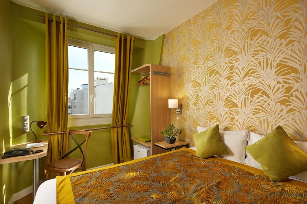 A hotel room with a bed, a desk, a chair, a window with curtains, a nightstand with a lamp, and a wall with a floral pattern.