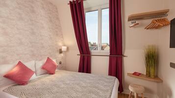 A bedroom with a bed, red curtains, a window with a view, a wooden shelf with clothes, and a small stool.