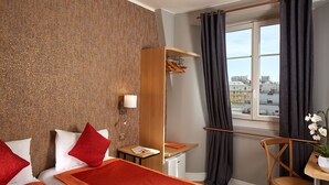 A hotel room with a bed, a desk, a chair, a small table with a plant, a window with curtains, and a view of buildings. - Hôtel Villa Sorel (Boulogne-Billancourt)