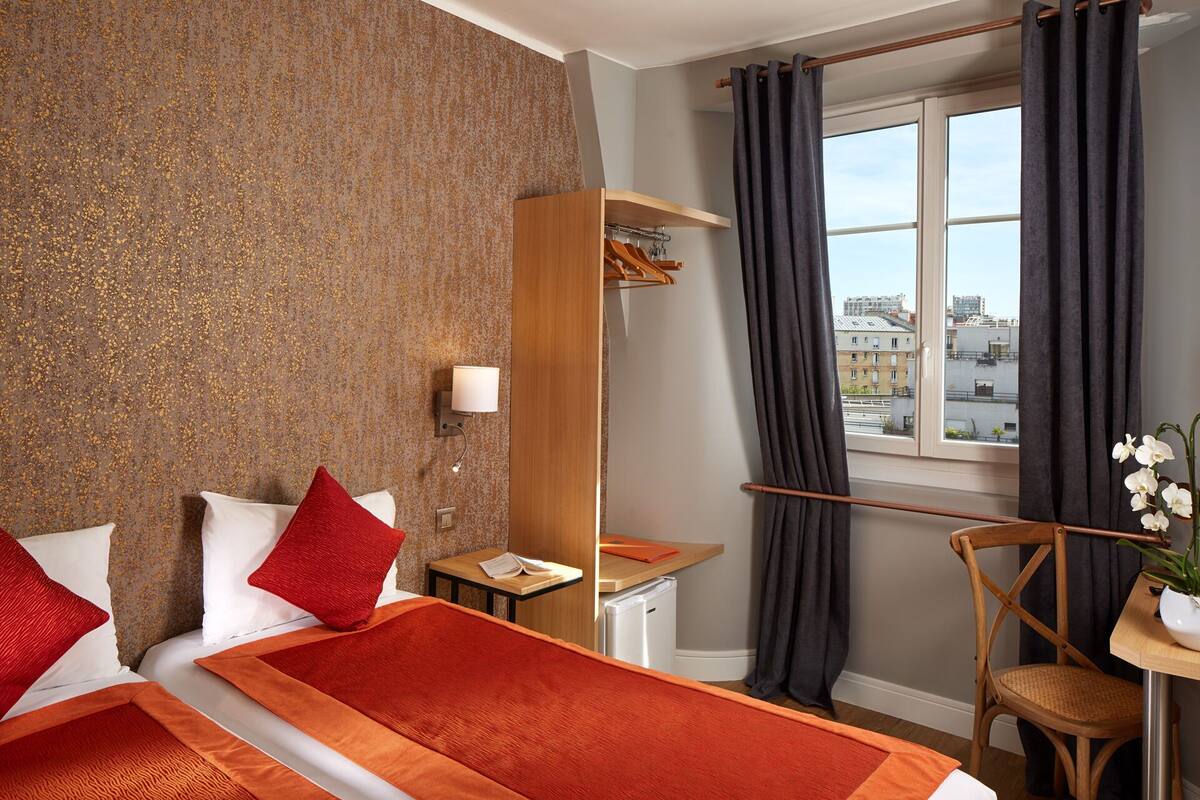A hotel room with a bed, a desk, a chair, a small table with a plant, a window with curtains, and a view of buildings.
