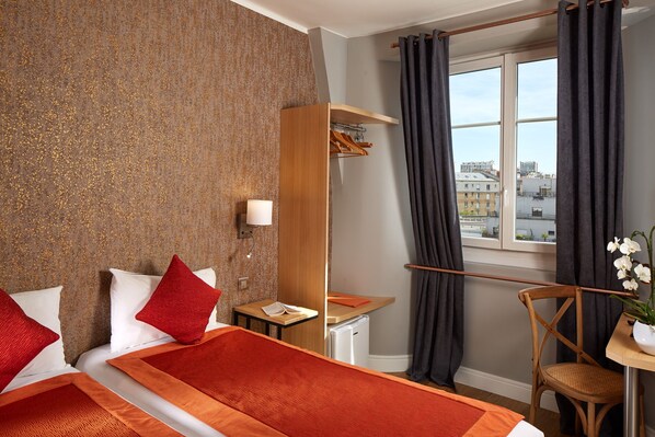 A hotel room with a bed, a desk, a chair, a small table with a plant, a window with curtains, and a view of buildings. - Hôtel Villa Sorel (Boulogne-Billancourt)