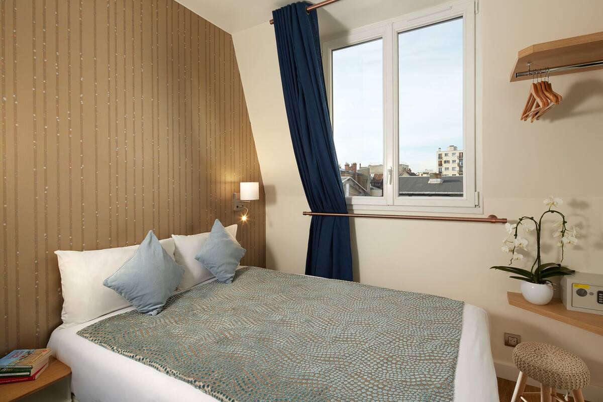 A bedroom with a bed, pillows, a window with curtains, a nightstand, a lamp, a plant, and a small stool.