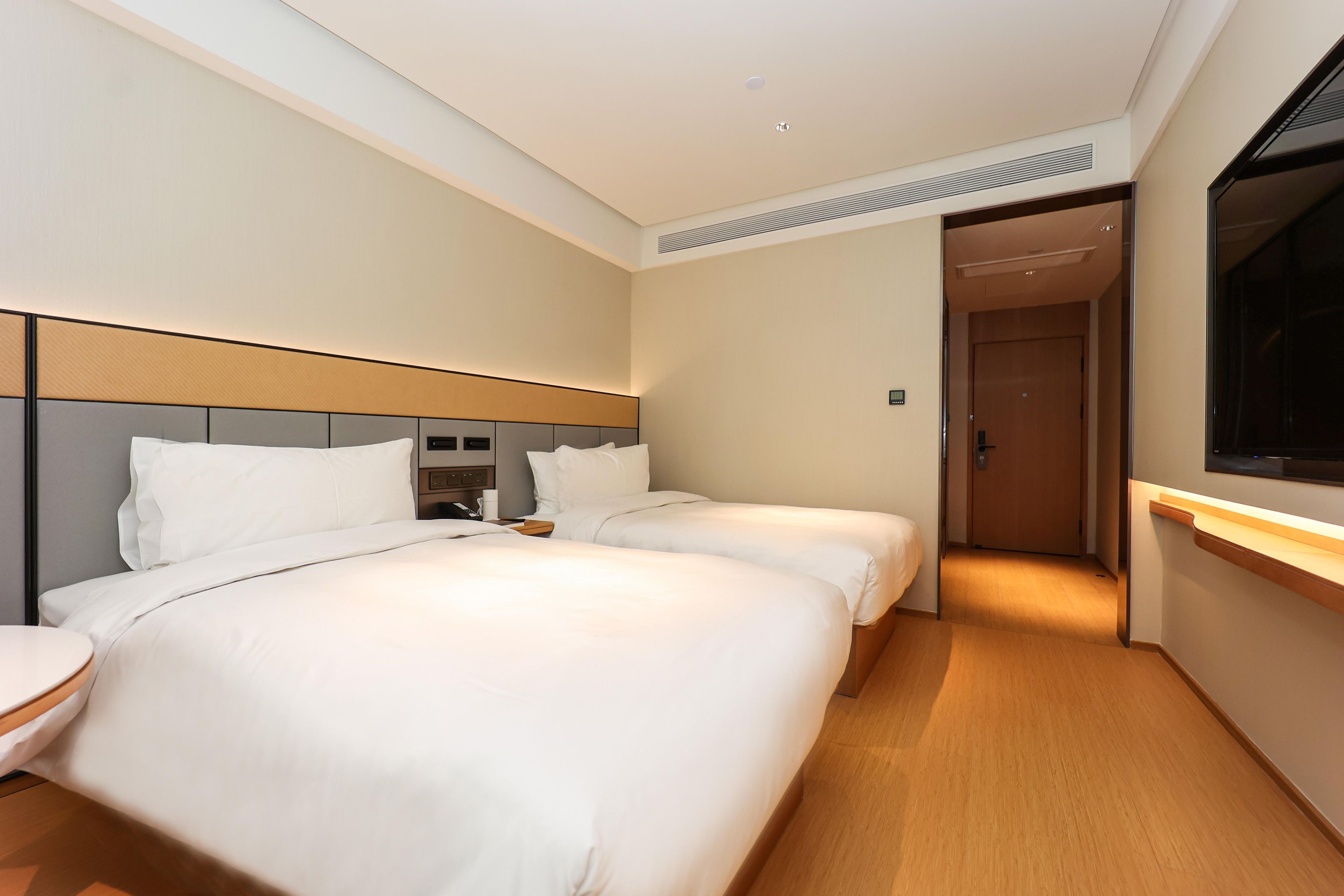 A hotel room with two beds, a bedside table, a TV, and a door leading to another room.