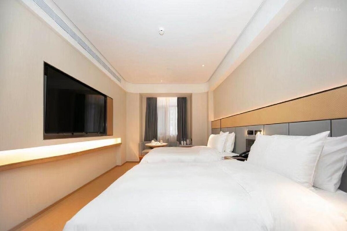A hotel room with two beds, a flat-screen TV mounted on the wall, a bedside table, and a window with curtains.