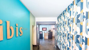 A hallway with a reception desk, a blue wall with the word 'bis' partially visible, and a patterned wall on the right. - ibis Esch Belval (Esch-sur-Alzette)