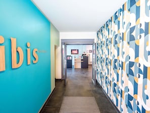 A hallway with a reception desk, a blue wall with the word 'bis' partially visible, and a patterned wall on the right. - ibis Esch Belval (Esch-sur-Alzette)