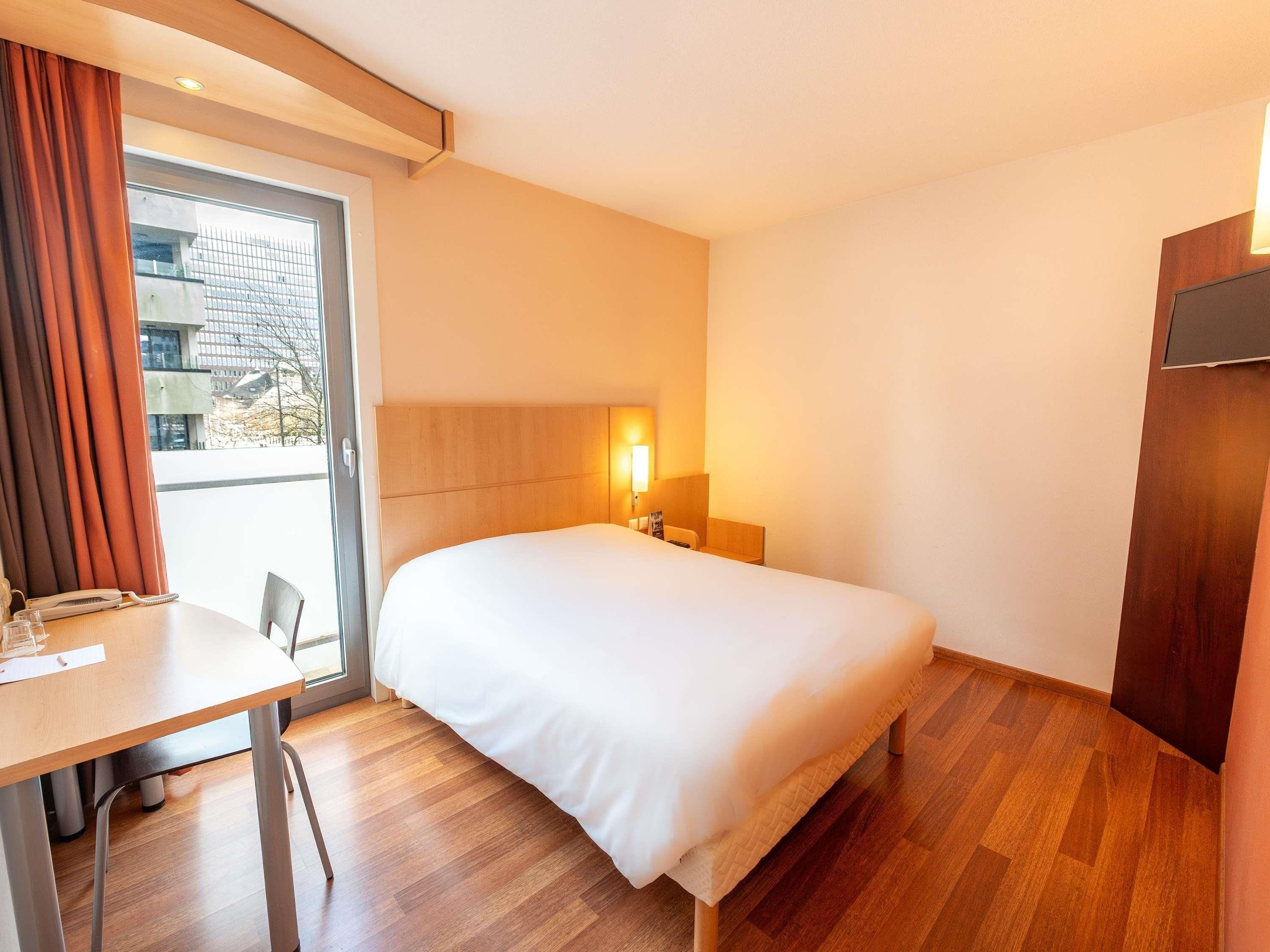 A hotel room with a bed, a desk, a chair, a lamp, and a view of the outside.
