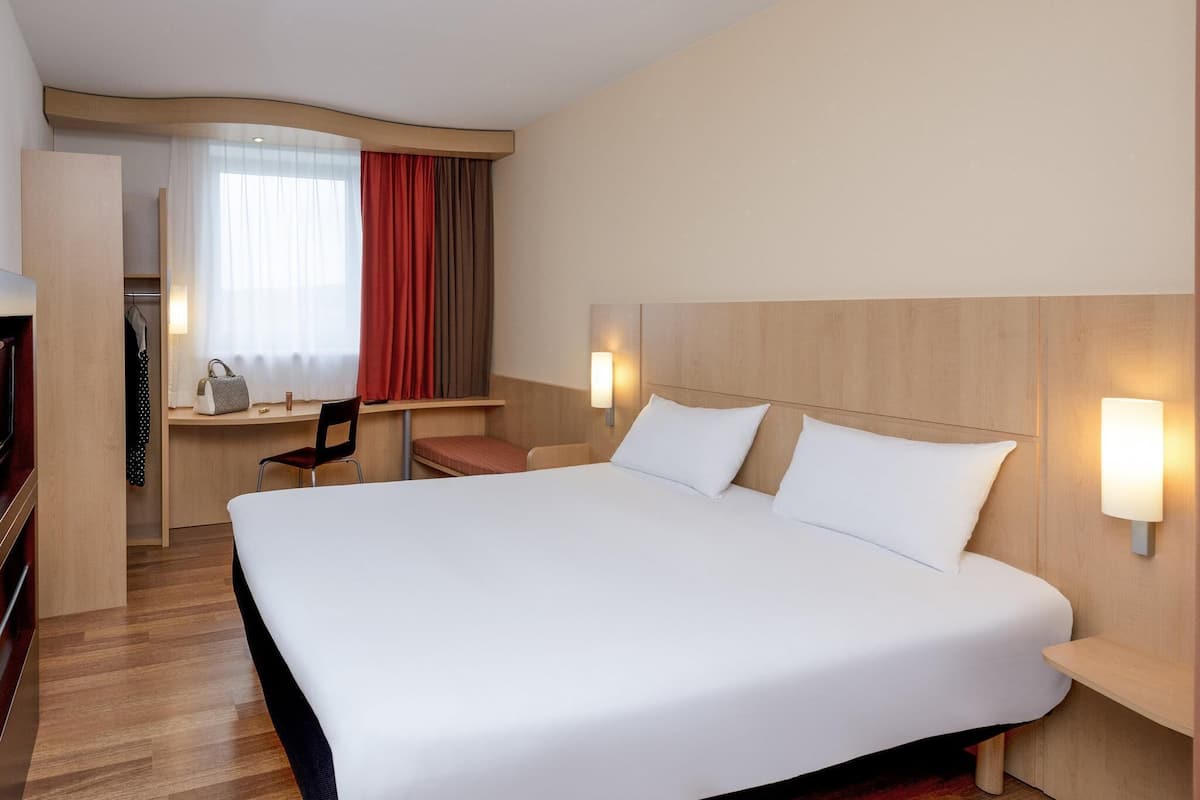 Hotellihuone, jossa on suuri sänky, työpöytä, tuoli, vaatekaappi ja ikkuna, jossa on verhot.