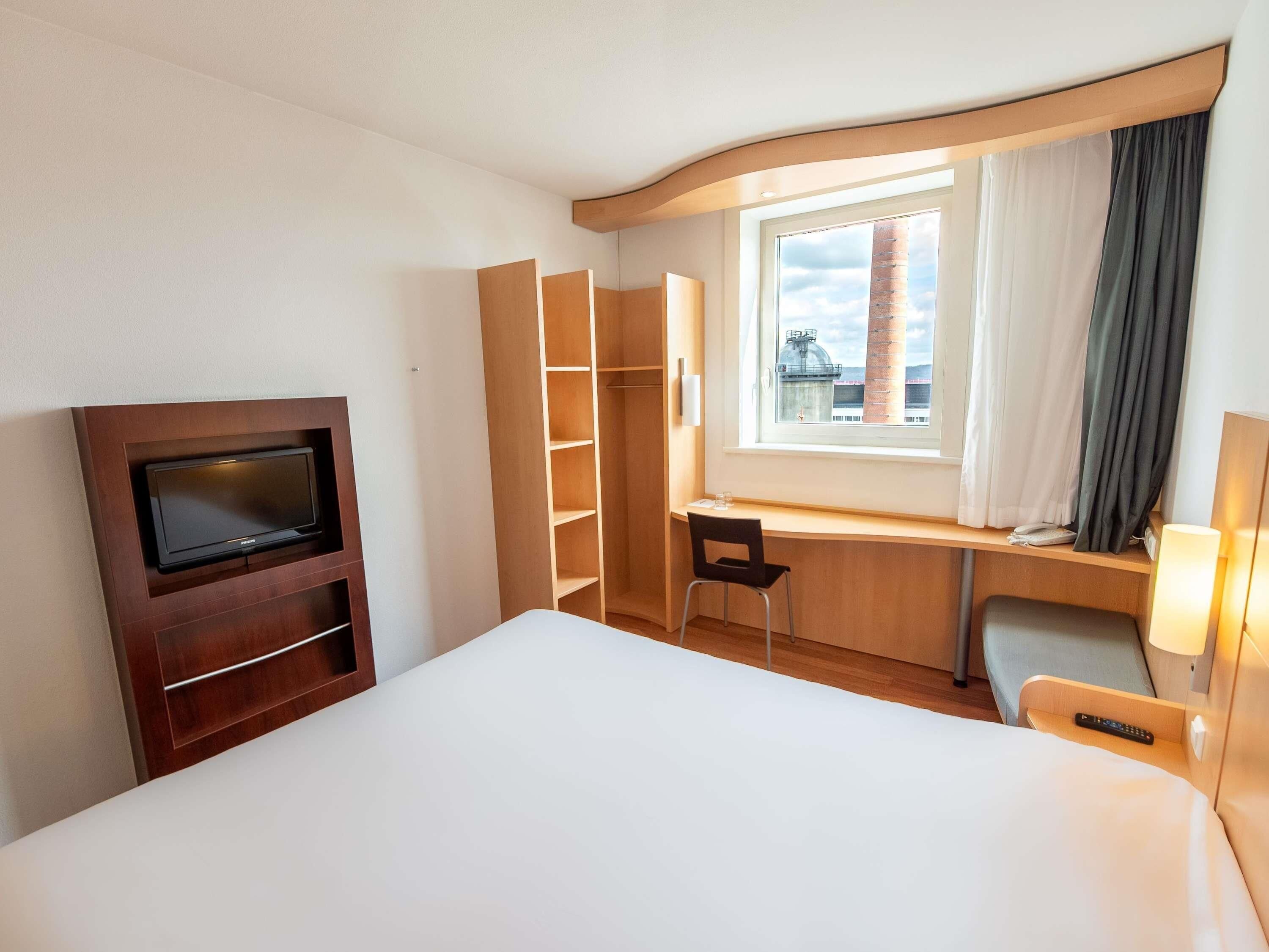 Habitación de hotel con cama, televisión, escritorio con silla, ventana con cortinas y armario.