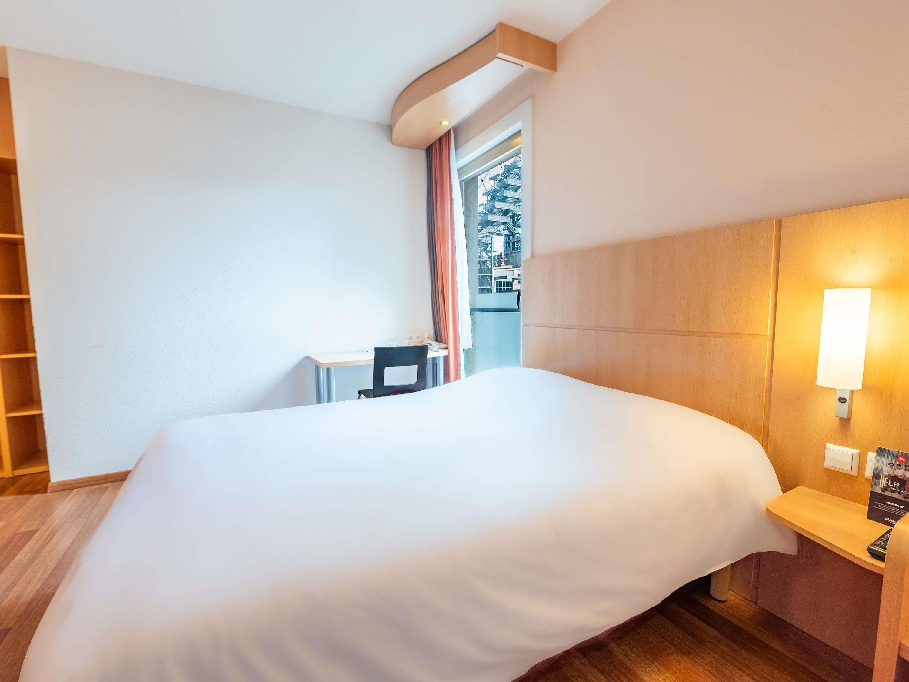 Hotelkamer met groot bed, bureau, stoel, nachtkastje en raam met gordijnen.