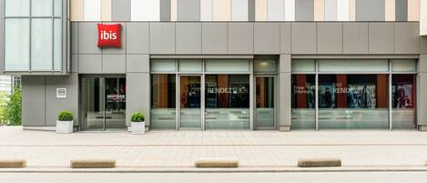 A modern building with a prominent red 'ibis' sign, large windows, and a clean, tiled pavement in front.
