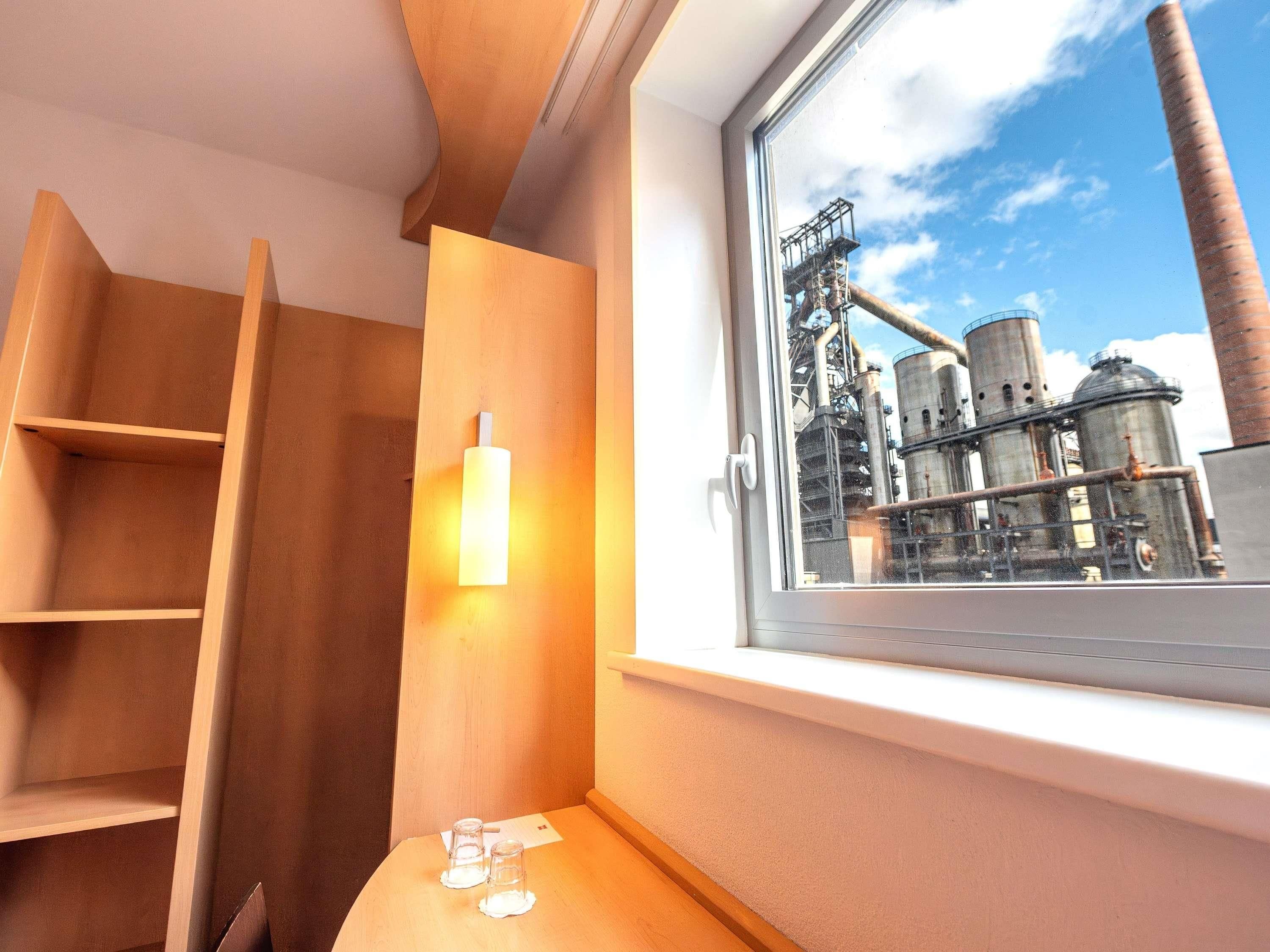 A room with a window overlooking an industrial area, wooden furniture, and a small table with glasses.