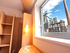 A room with a window overlooking an industrial area, wooden furniture, and a small table with glasses. - ibis Esch Belval (Esch-sur-Alzette)