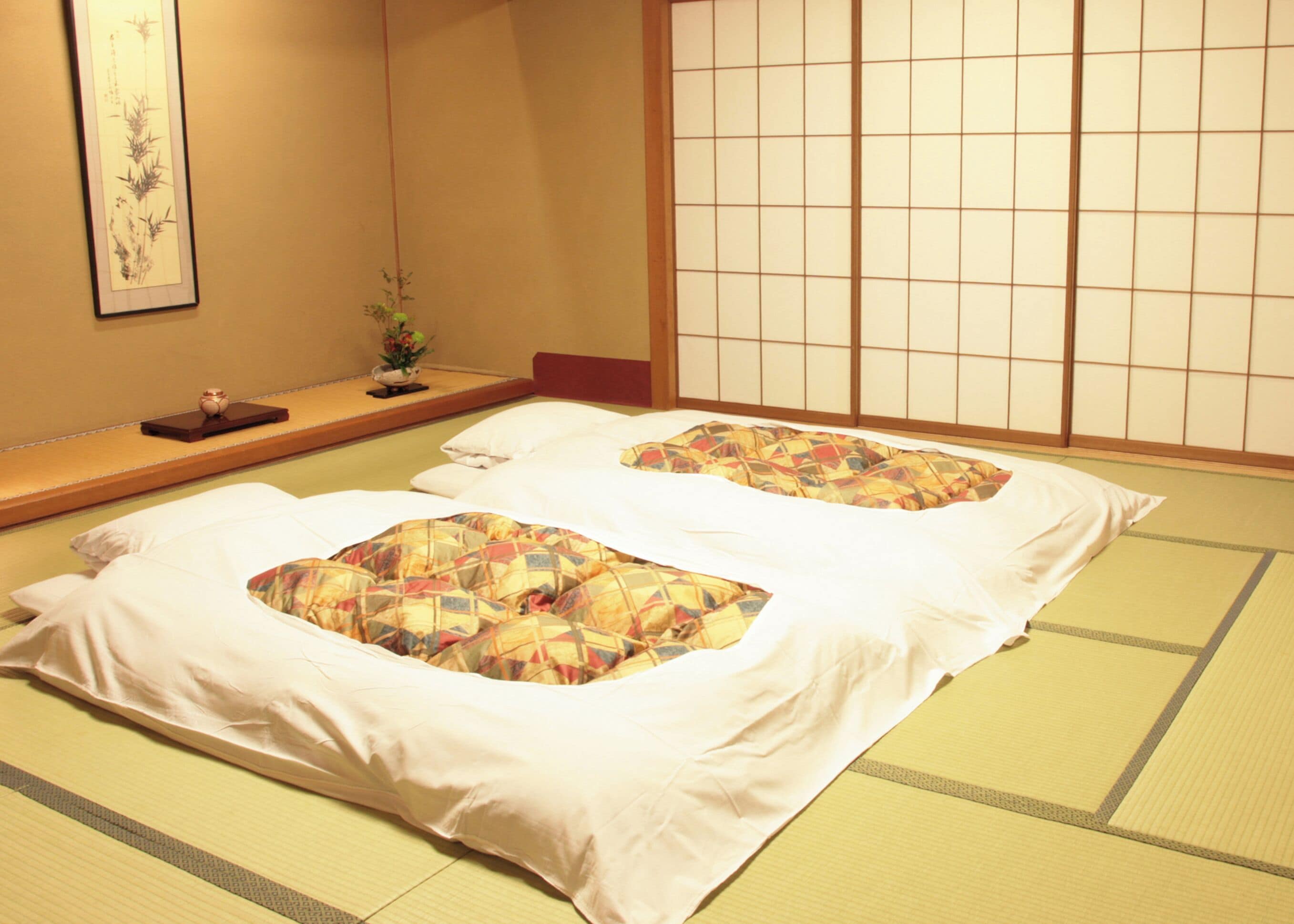 A traditional Japanese room with tatami flooring, a low wooden bed with white bedding, and a shoji screen in the background.