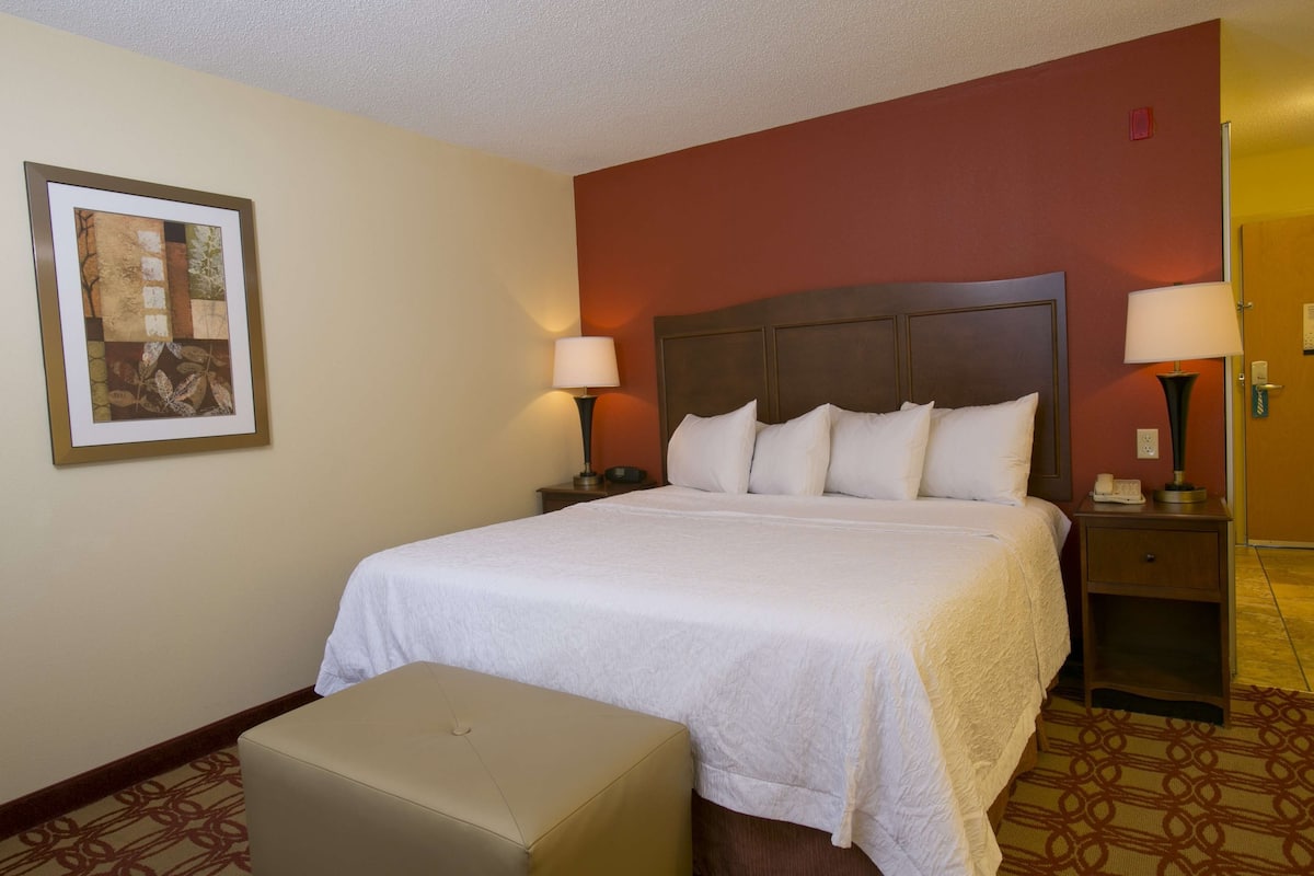 A hotel room with a large bed, two bedside tables with lamps, a small bench, and a framed picture on the wall.