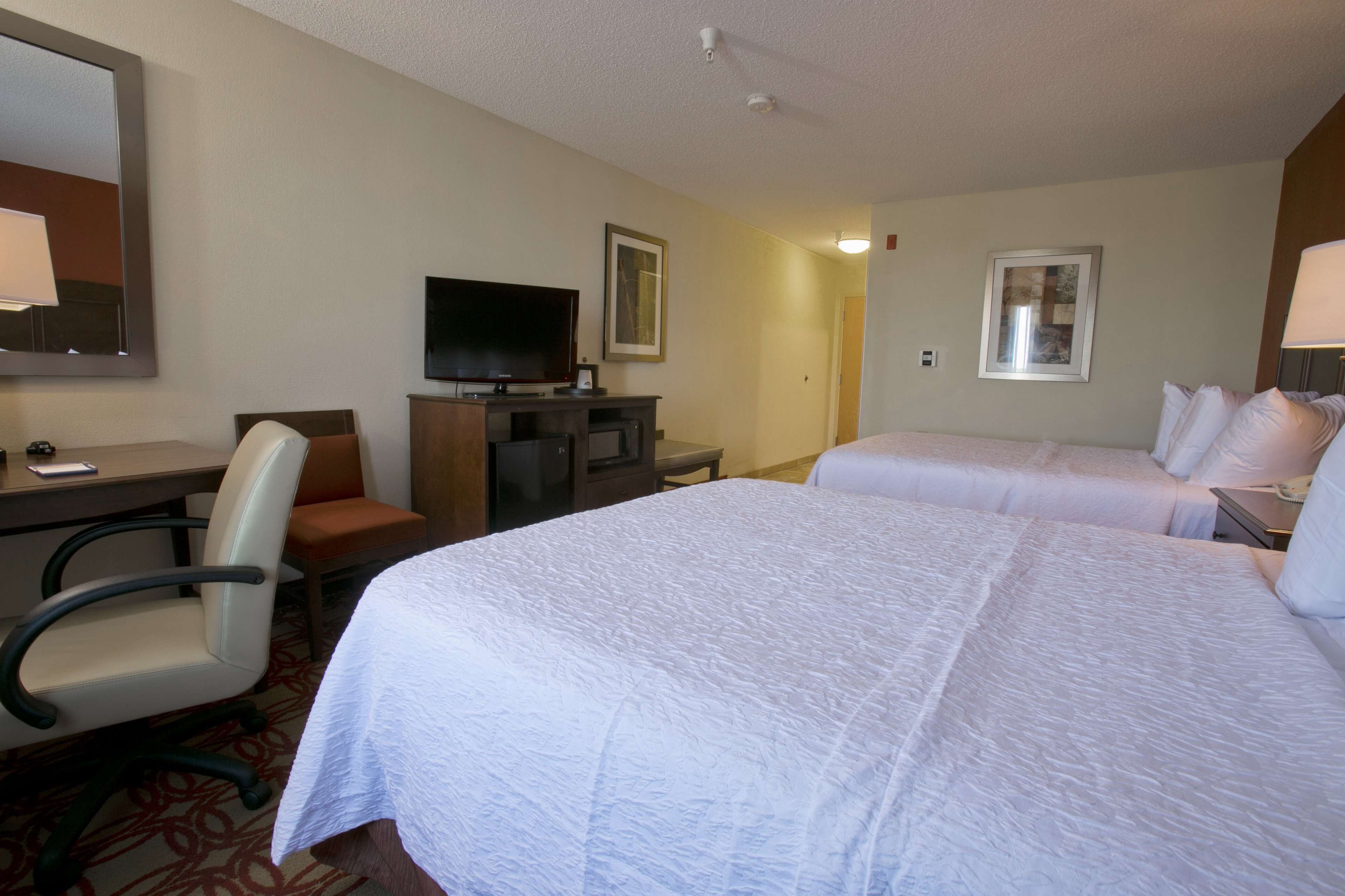 A hotel room with two beds, a desk with a chair, a television, and framed pictures on the walls.